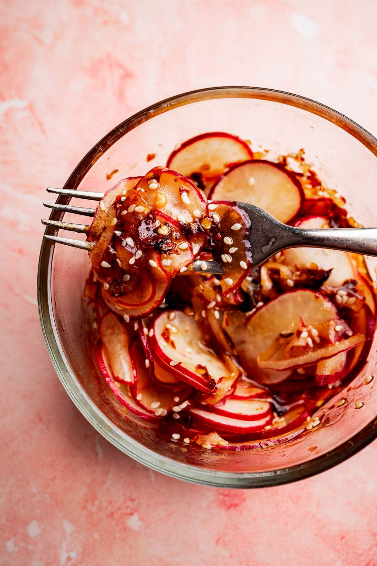A glass bowl of sliced radishes mixed with sesame seeds and sauce, part of a spicy radish recipe, with a fork lifting a portion on a pink surface.