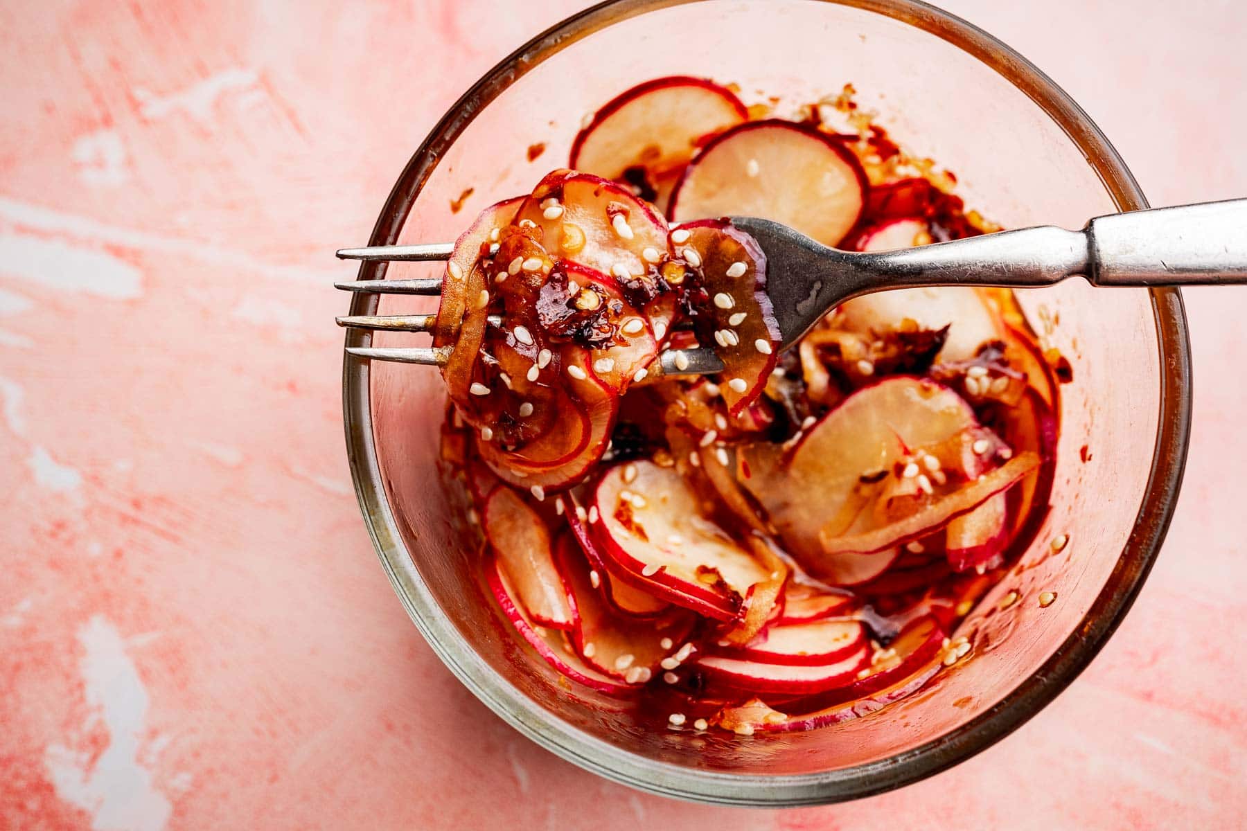 A fork holds thinly sliced radishes with sauce and sprinkled sesame seeds above a glass bowl on a pink surface, showcasing a vibrant spicy radish recipe.
