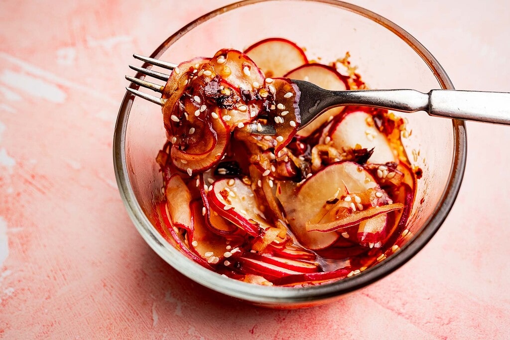 A glass bowl containing a spicy radish recipe: thinly sliced radishes mixed with sauce and sprinkled with sesame seeds, with a fork resting inside.