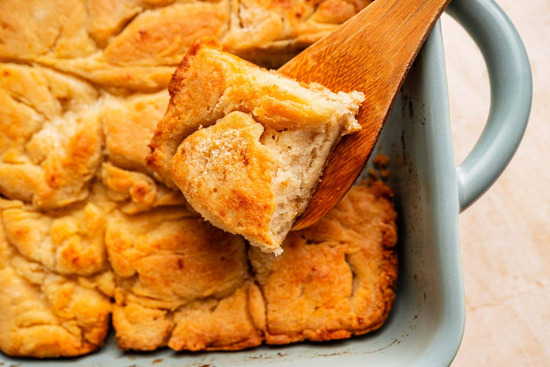 A close-up of golden-brown gluten free butter swim biscuits in a baking dish, with one biscuit being lifted out on a wooden spatula.