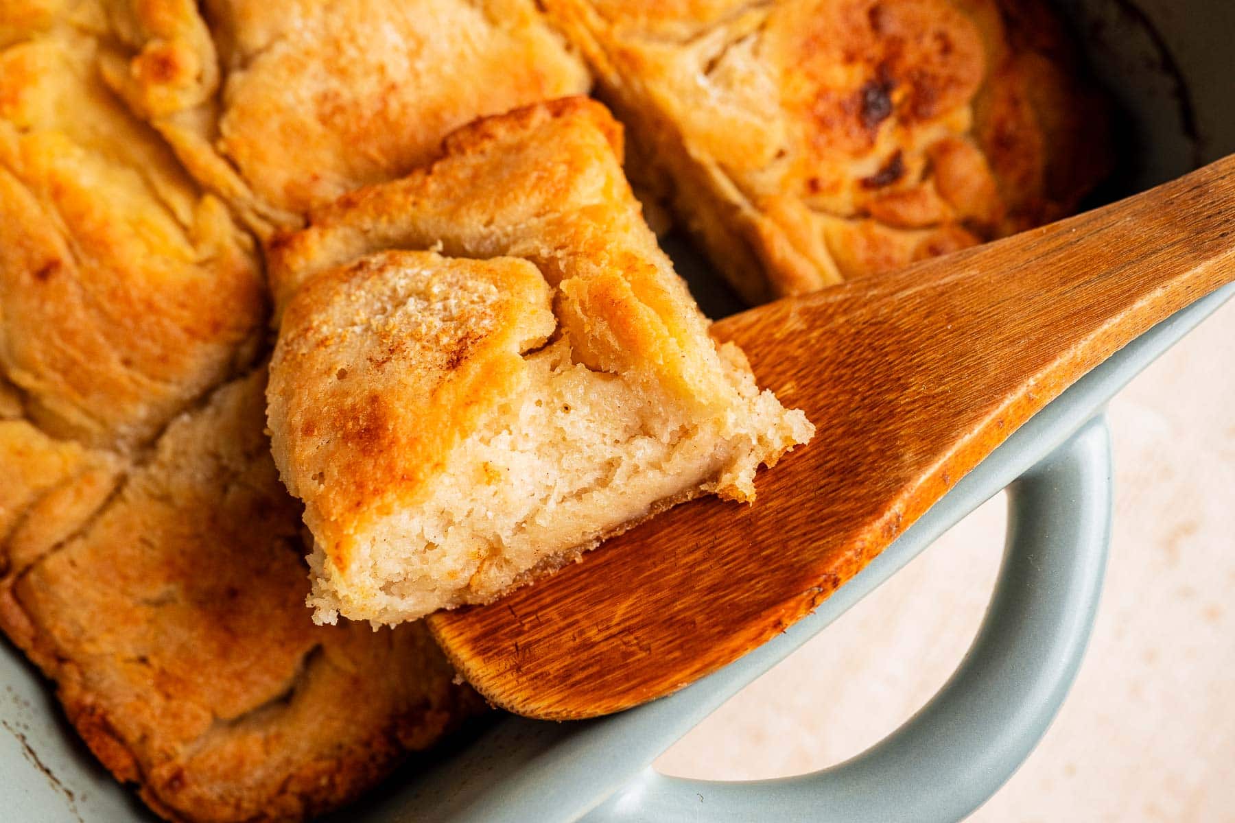 A close-up of a golden, flaky gluten free butter swim biscuit being lifted from a baking dish with a wooden spatula.