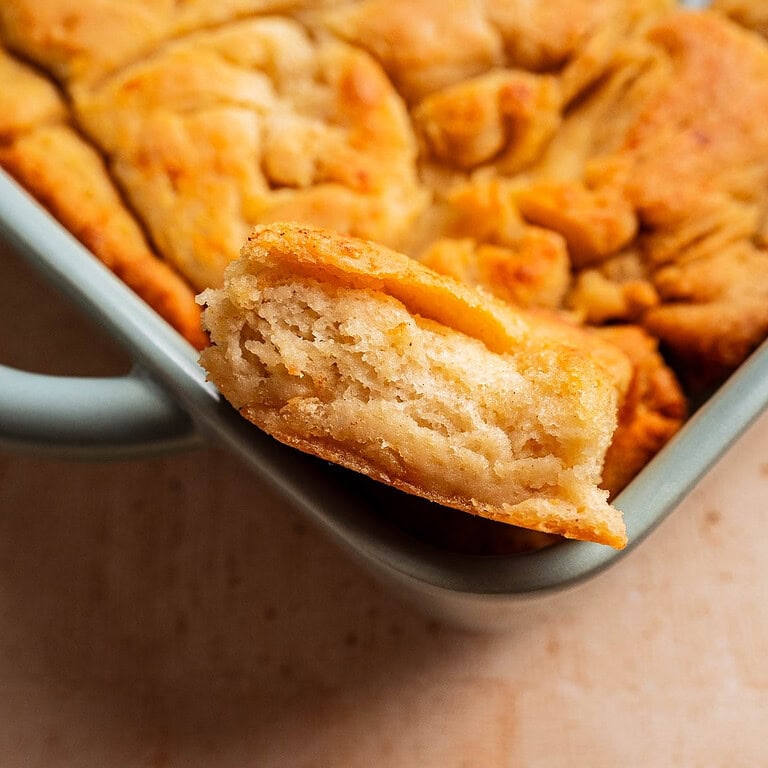A close-up of a piece of pull-apart bread with a flaky, layered texture, reminiscent of gluten free butter swim biscuits, held above a baking dish containing the rest of the bread.