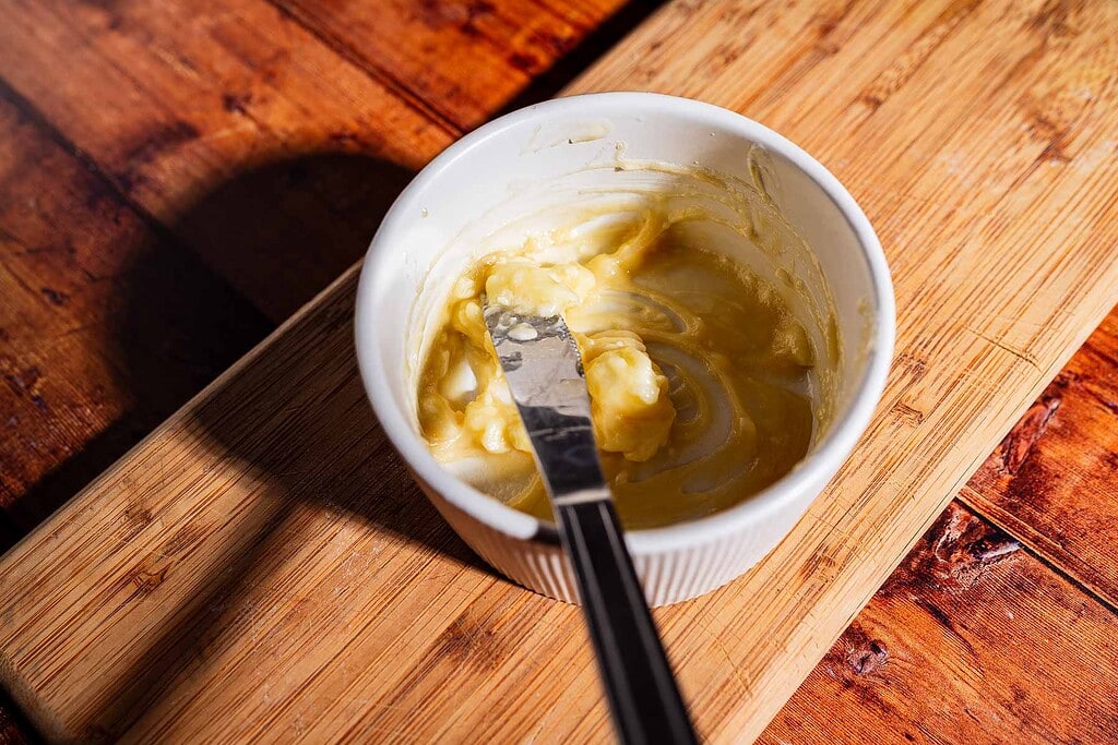 A small white ramekin with a butter mixture for kimchi grilled cheese and a butter knife sits on a wooden cutting board.