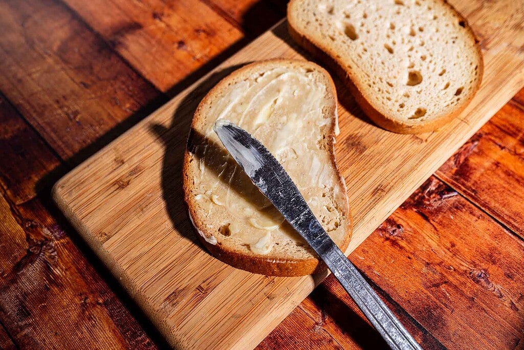 A slice of bread is being spread with butter using a knife on a wooden cutting board, with another slice of bread nearby—getting prepped for a delicious kimchi grilled cheese.