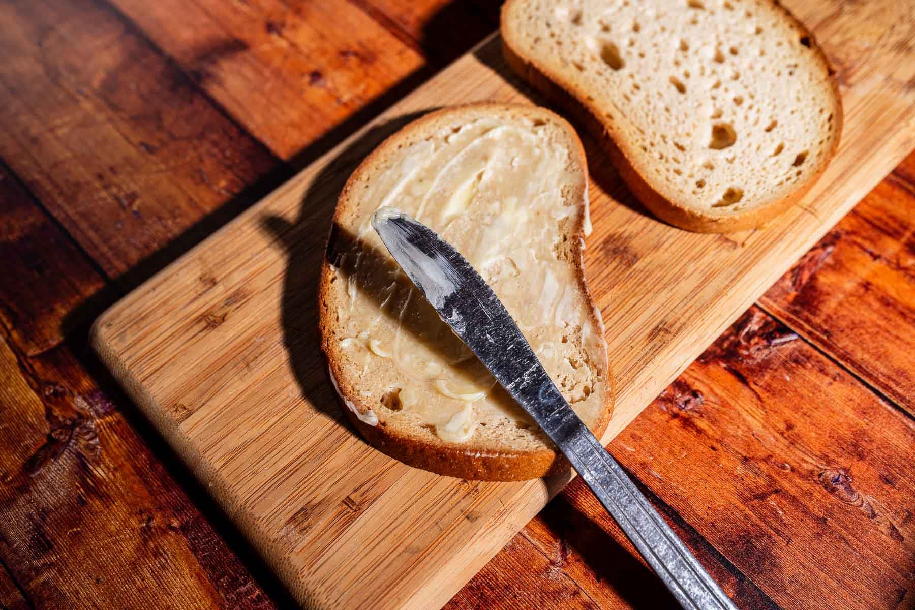 A slice of bread is being spread with butter using a knife on a wooden cutting board, with another slice of bread nearby—getting prepped for a delicious kimchi grilled cheese.