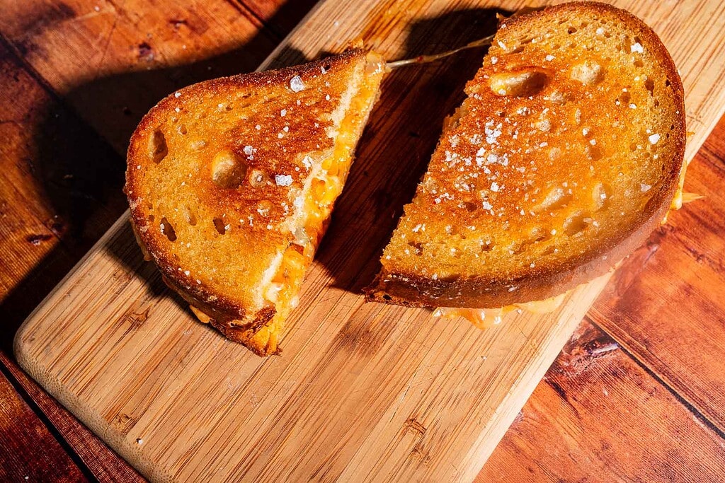 A kimchi grilled cheese sandwich, cut in half, sits on a wooden cutting board on a wooden table.