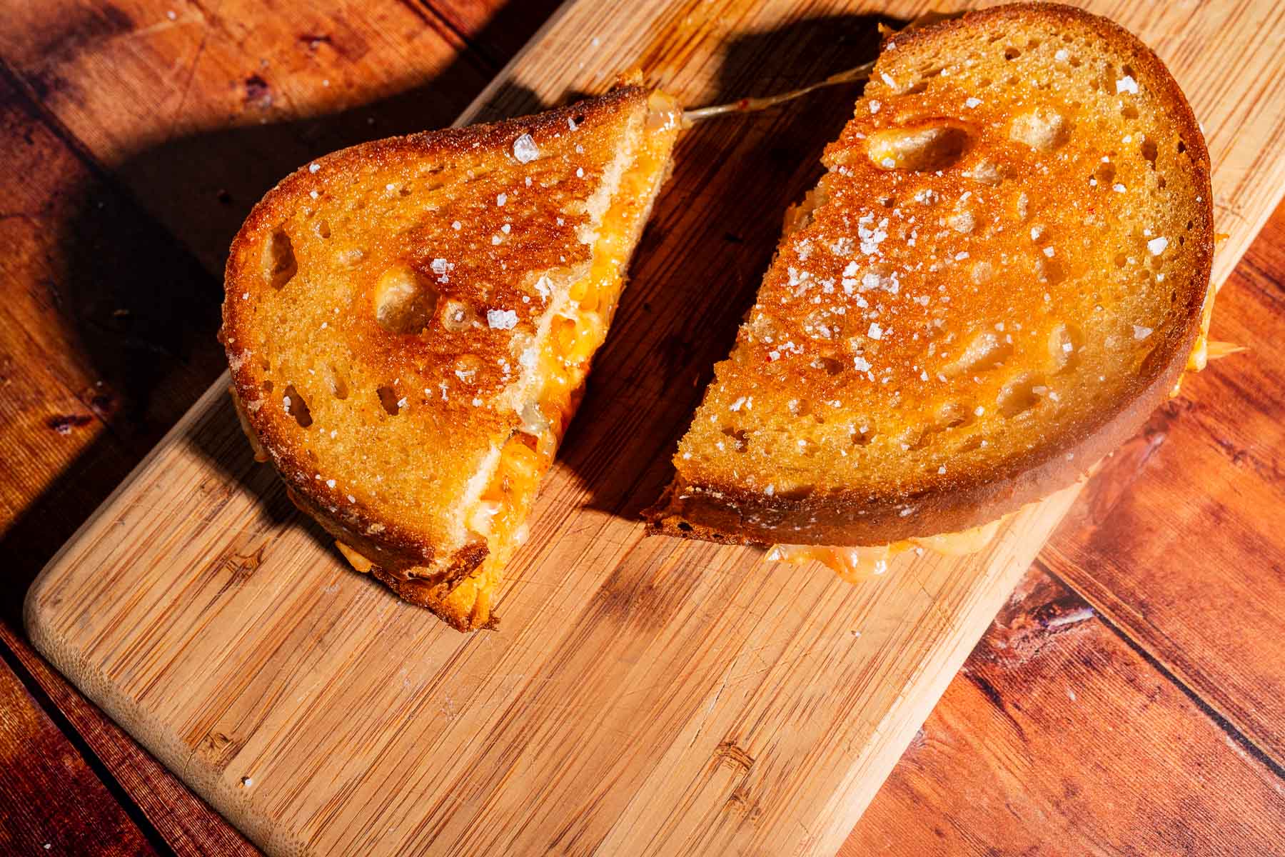 A kimchi grilled cheese sandwich, cut in half, sits on a wooden cutting board on a wooden table.