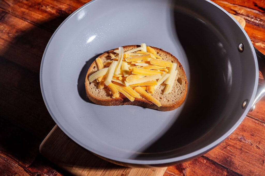 A slice of bread topped with shredded cheese sits in a gray frying pan on a wooden surface, ready to become a delicious kimchi grilled cheese.