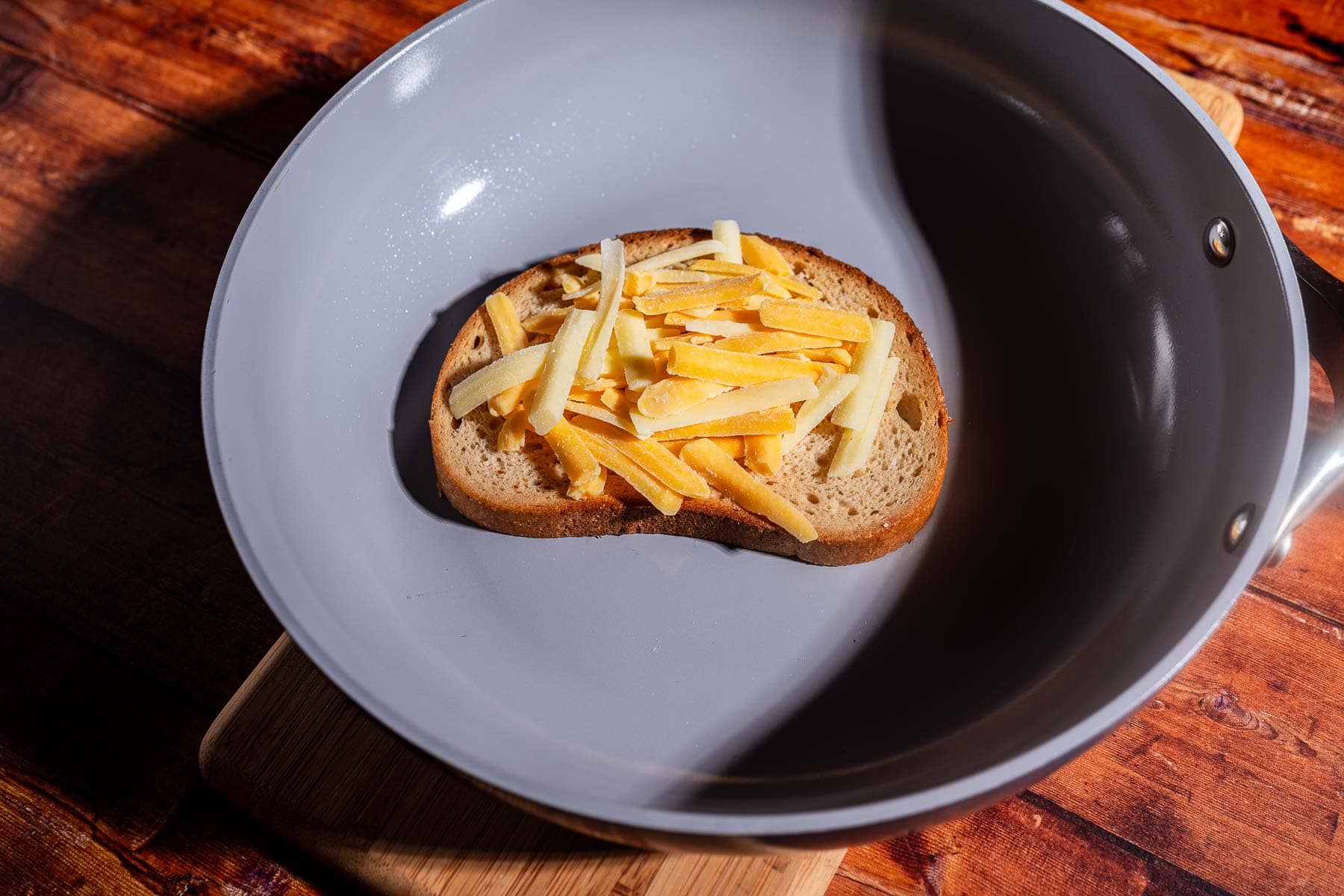 A slice of bread topped with shredded cheese sits in a gray frying pan on a wooden surface, ready to become a delicious kimchi grilled cheese.