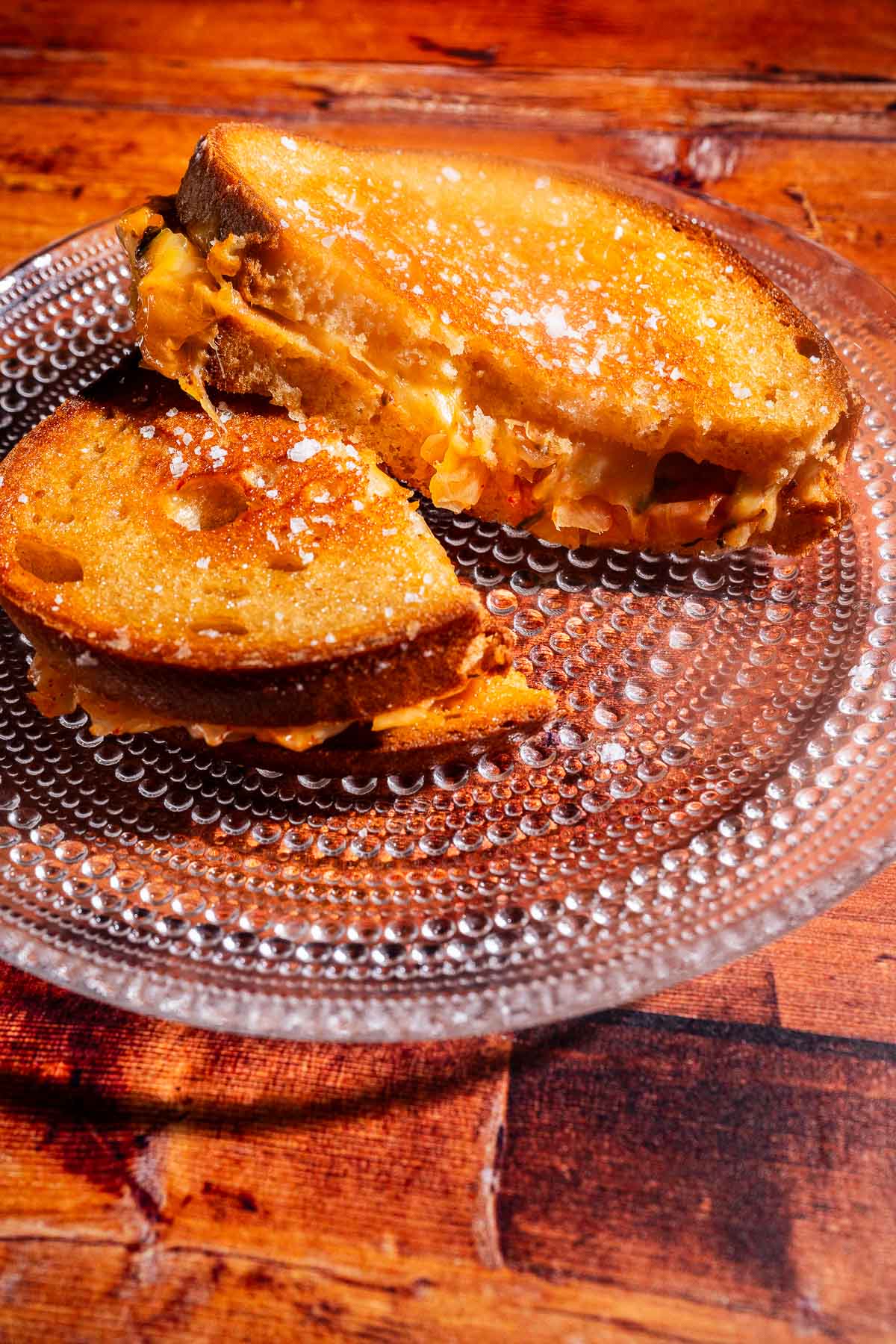 A kimchi grilled cheese sandwich with a golden crust is cut in half and served on a clear glass plate on a wooden table.