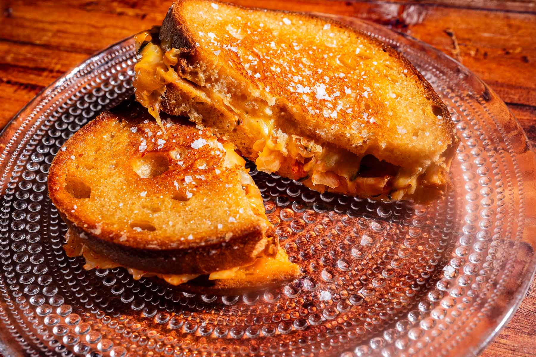 A kimchi grilled cheese sandwich cut in half sits on a clear textured glass plate, with a dusting of salt on the golden toasted bread.