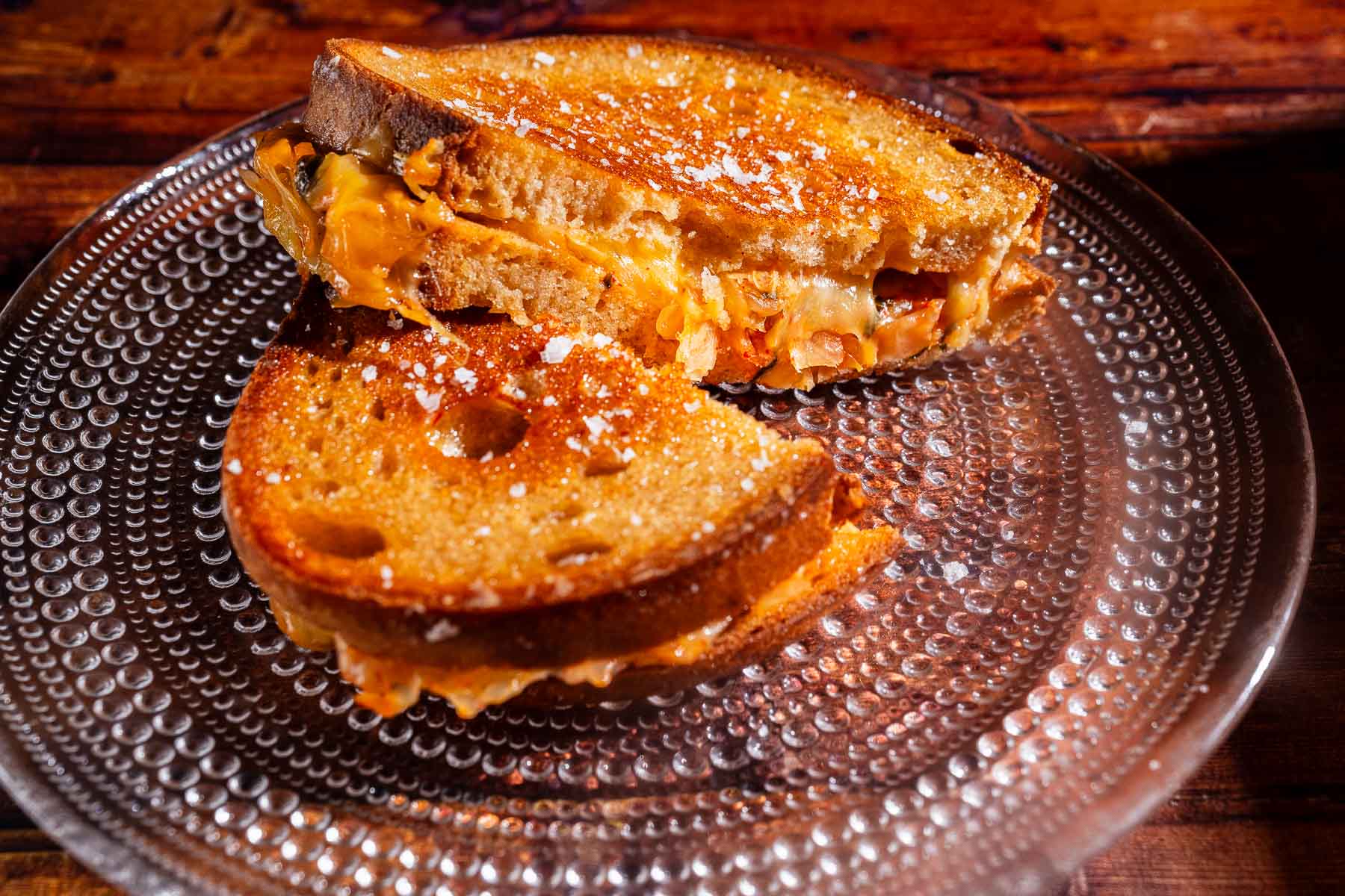 A kimchi grilled cheese sandwich cut in half, with melted cheese and tangy kimchi oozing between two slices of toasted bread, served on a textured glass plate.
