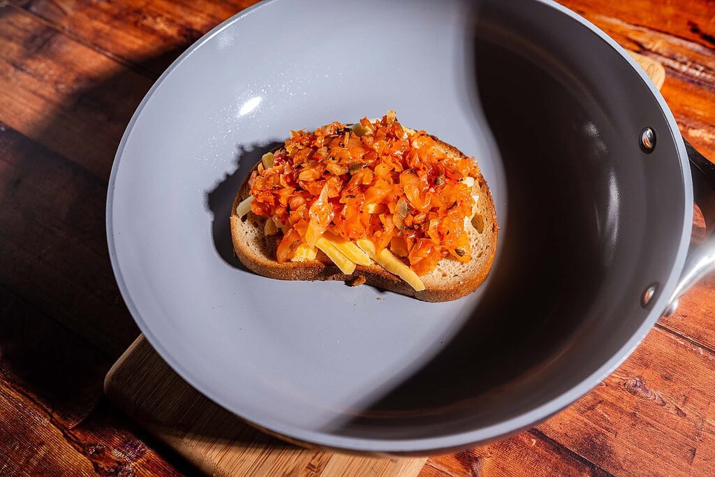 A slice of bread topped with sautéed vegetables and melty kimchi grilled cheese sits in a gray frying pan on a wooden surface.