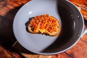 A slice of bread topped with sautéed vegetables and melty kimchi grilled cheese sits in a gray frying pan on a wooden surface.