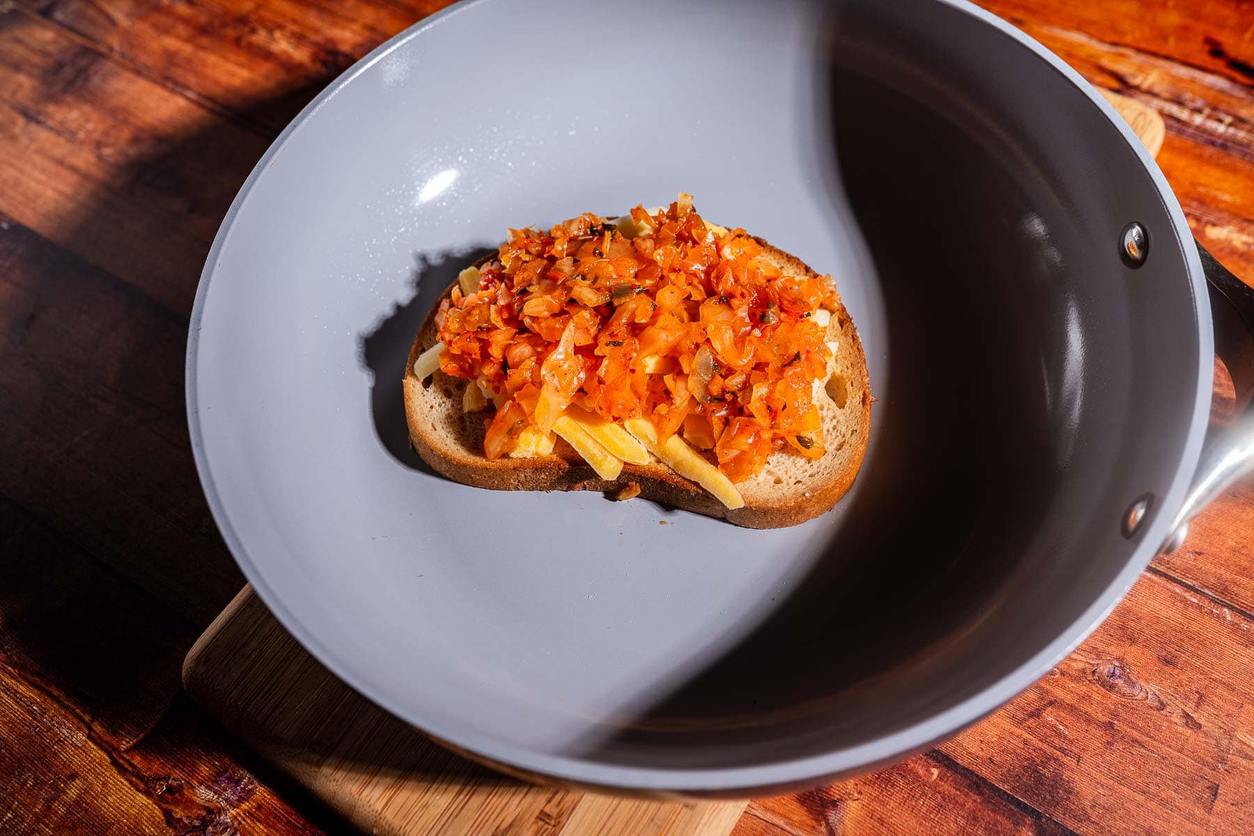 A slice of bread topped with sautéed vegetables and melty kimchi grilled cheese sits in a gray frying pan on a wooden surface.