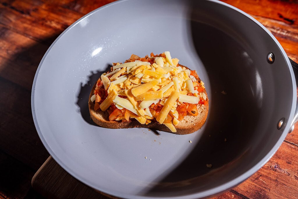 A slice of bread topped with grated cheese, chopped vegetables, and tangy kimchi sits in a gray nonstick frying pan on a wooden surface, creating the perfect kimchi grilled cheese.