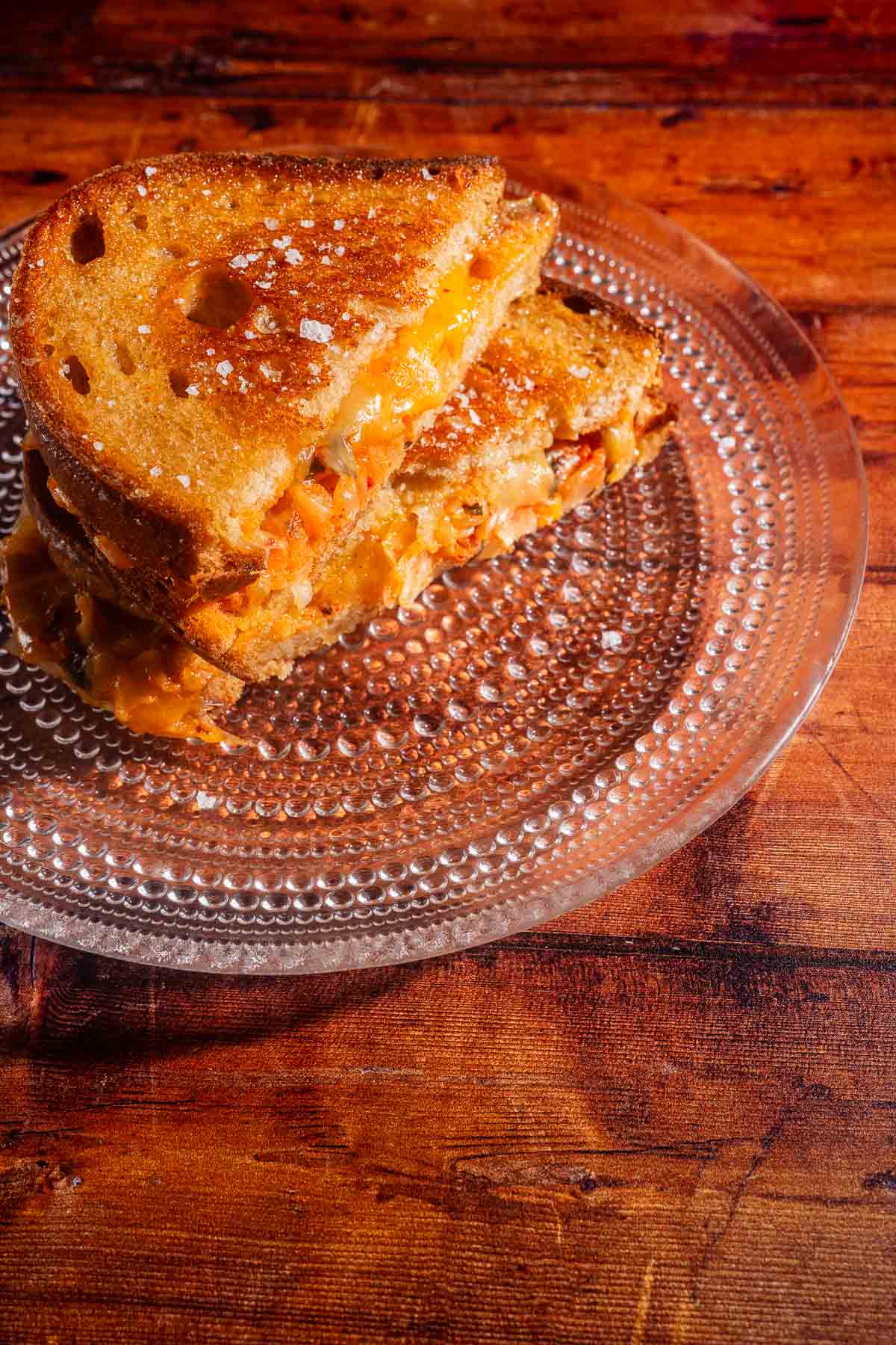 A kimchi grilled cheese sandwich cut in half, with melted cheese and kimchi visible, sits on a clear glass plate on a wooden table.
