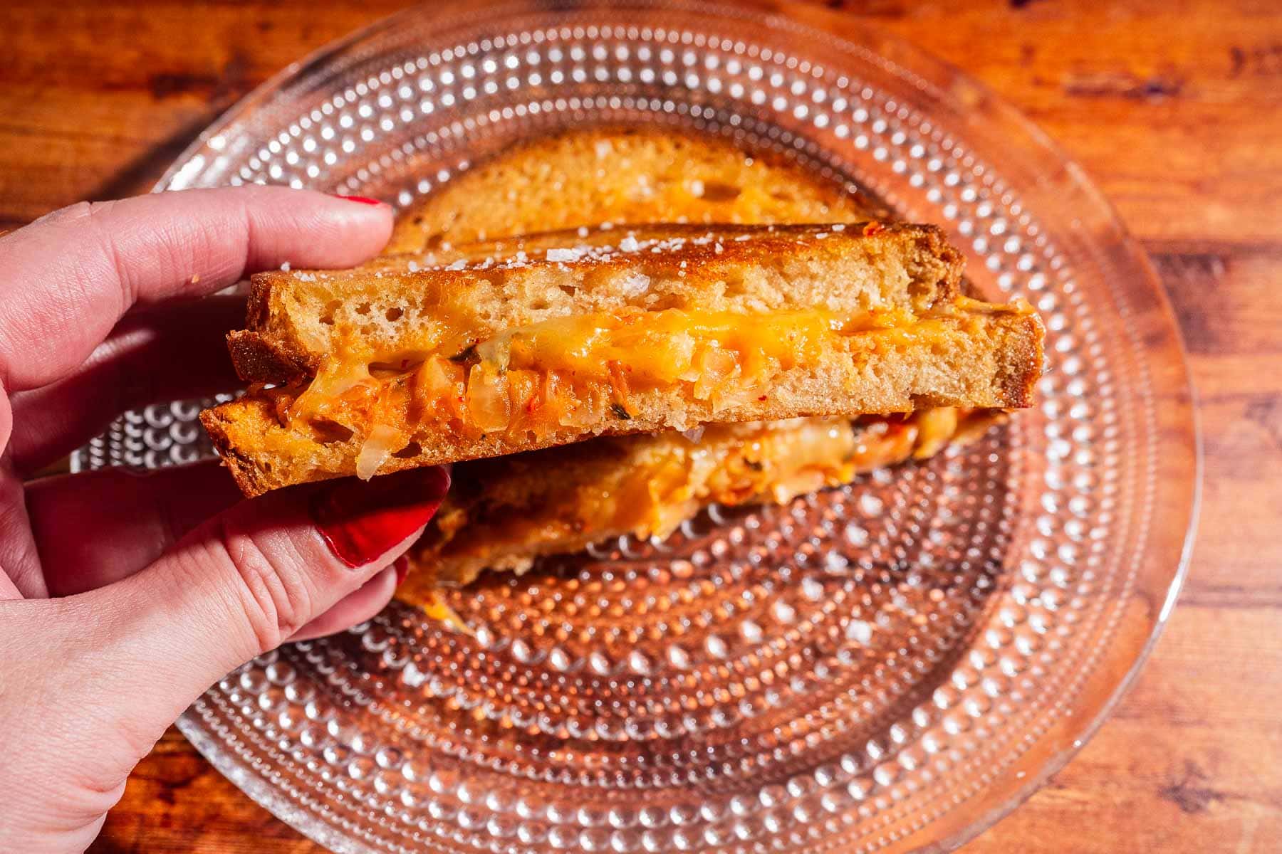 A hand holding half of a kimchi grilled cheese sandwich with melted cheese, on a glass plate atop a wooden table.