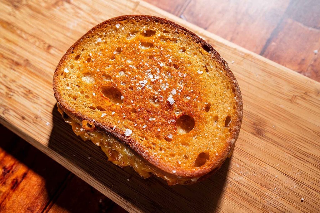 A kimchi grilled cheese sandwich with golden brown, crispy bread and a sprinkling of salt sits on a wooden cutting board.