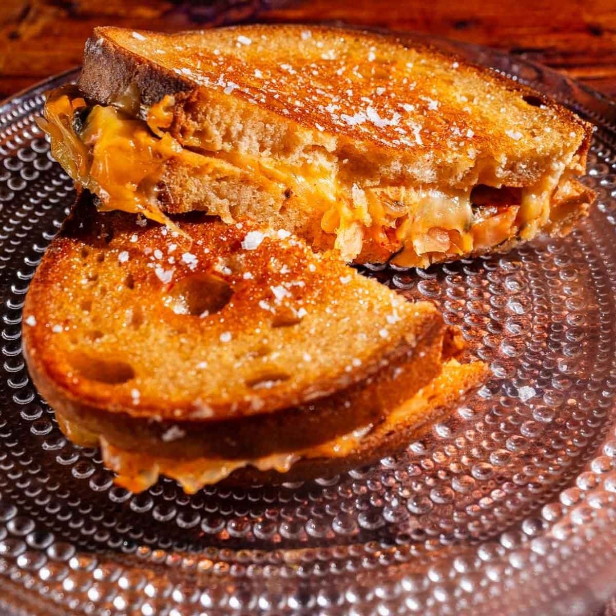 A kimchi grilled cheese sandwich with melted cheese is cut in half and served on a clear glass plate.