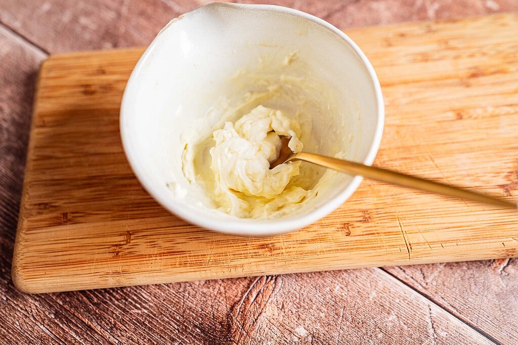 A small white bowl with a gold spoon contains a creamy jalapeno popper grilled cheese mixture, placed on a wooden cutting board.