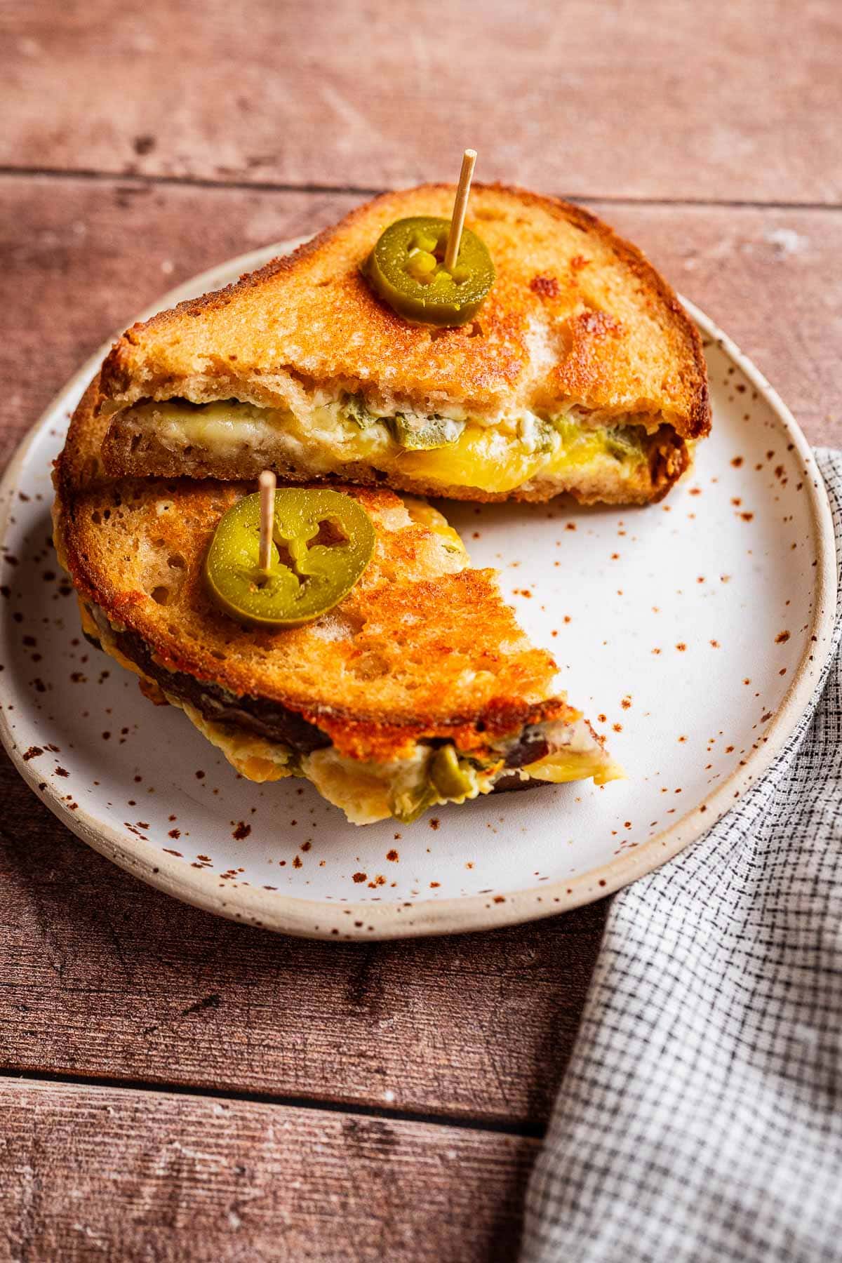 A jalapeno popper grilled cheese sandwich cut in half, topped with jalapeño slices, served on a speckled plate with a toothpick in each half.