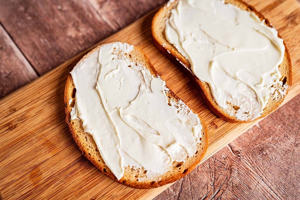 Two slices of bread with cream cheese spread on top, perfect for making a delicious jalapeno popper grilled cheese, placed on a wooden cutting board.