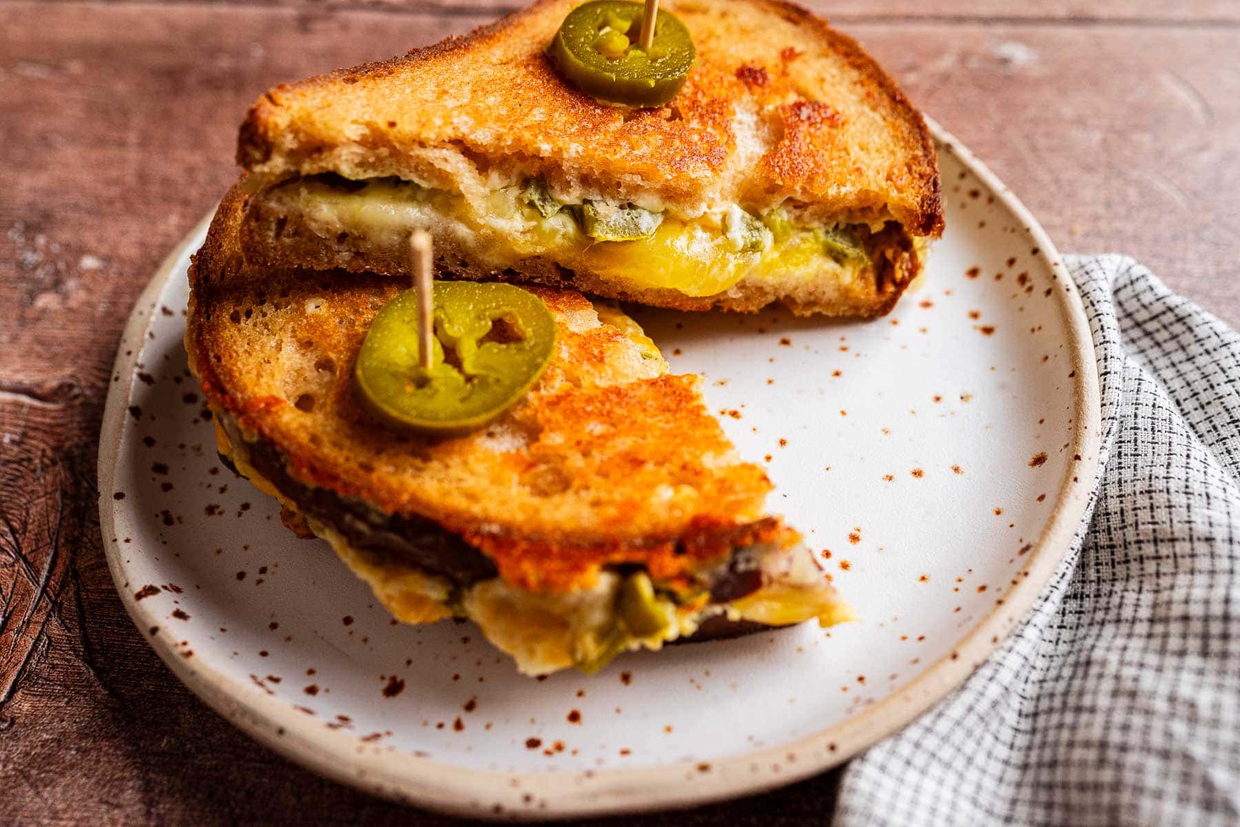 A jalapeno popper grilled cheese sandwich with spicy jalapeño slices, cut in half and served on a white plate with a toothpick, next to a checkered napkin.