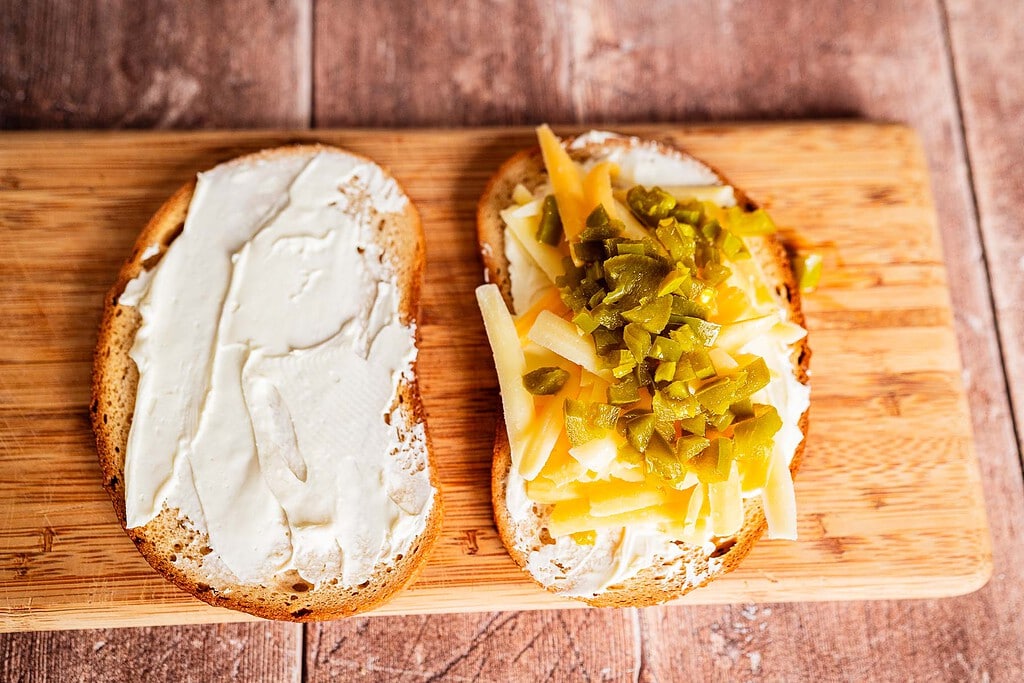 Two slices of bread on a wooden board; one with cream cheese, the other topped with cream cheese, shredded cheese, chopped green peppers—perfect for assembling a jalapeno popper grilled cheese.