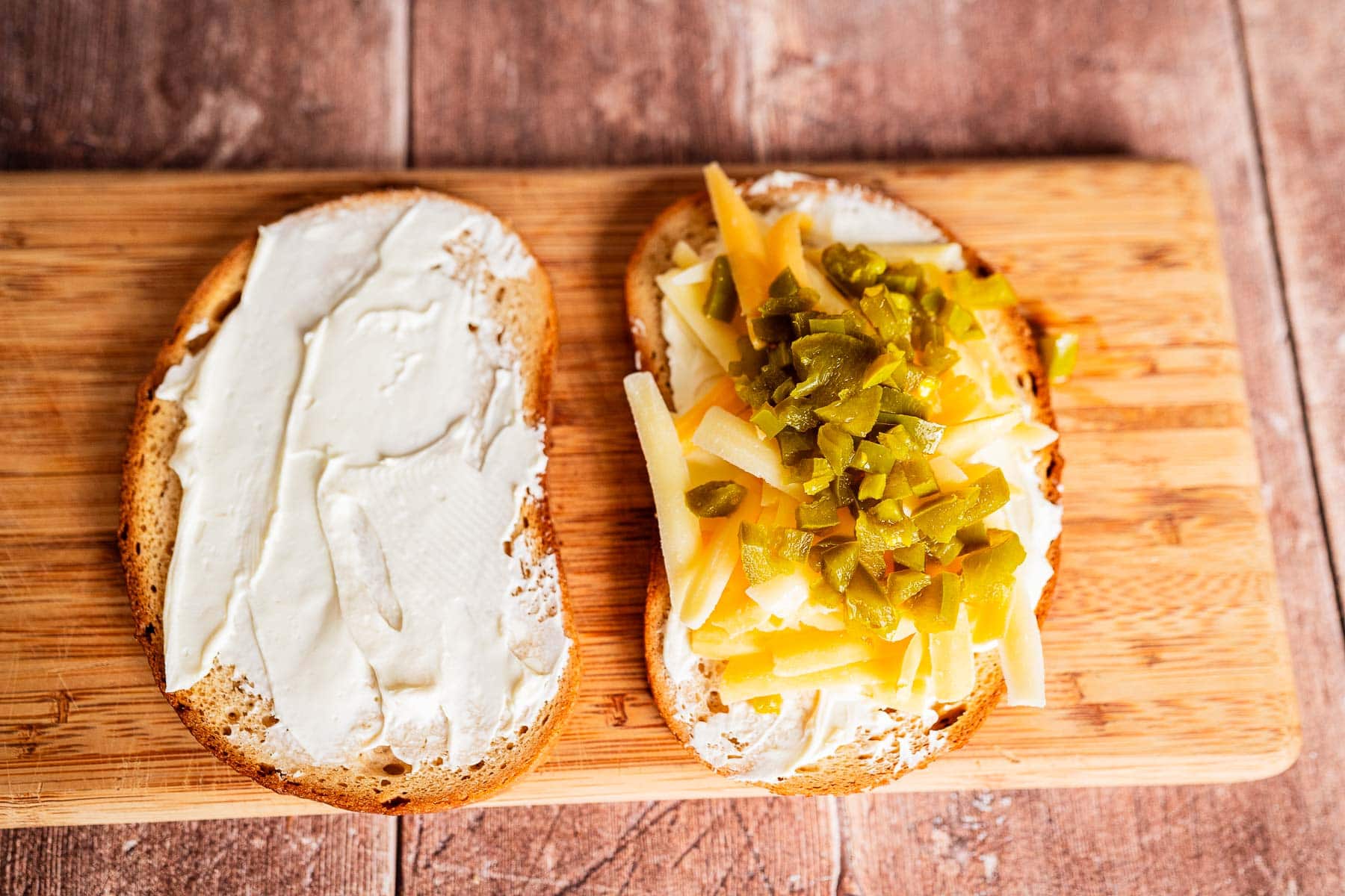 Two slices of bread on a wooden board; one with cream cheese, the other topped with cream cheese, shredded cheese, chopped green peppers—perfect for assembling a jalapeno popper grilled cheese.