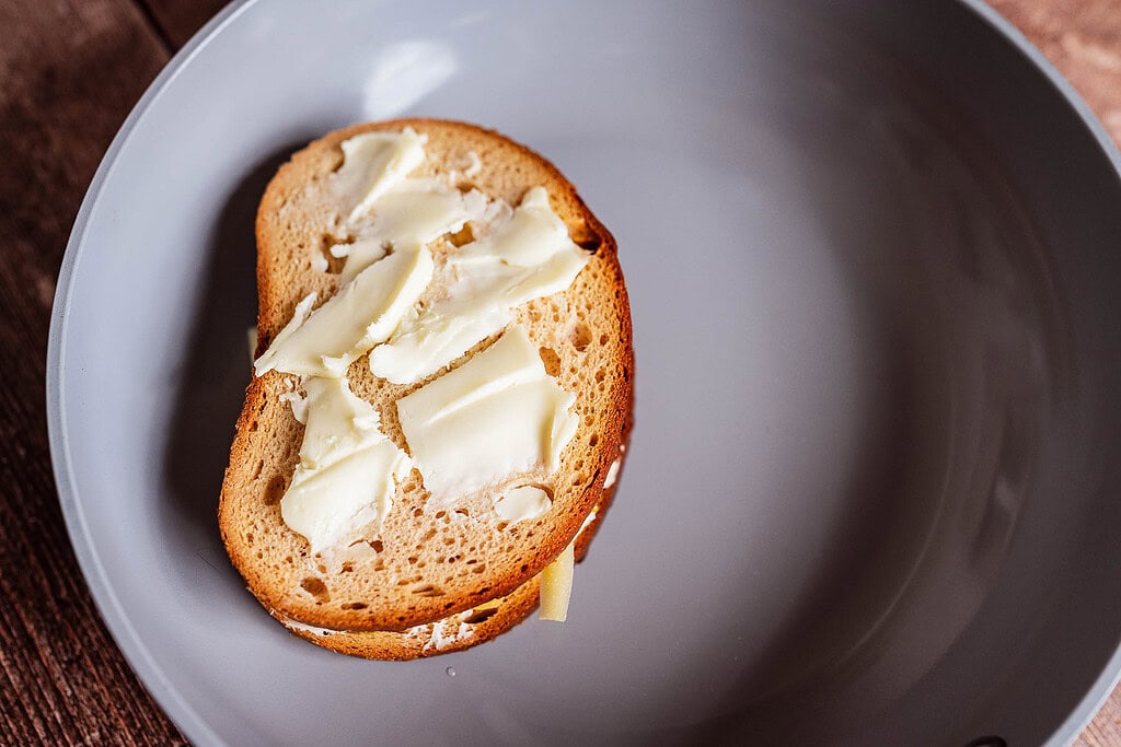 A slice of bread with several pieces of butter on top sits in a gray frying pan, ready to become a jalapeno popper grilled cheese masterpiece.
