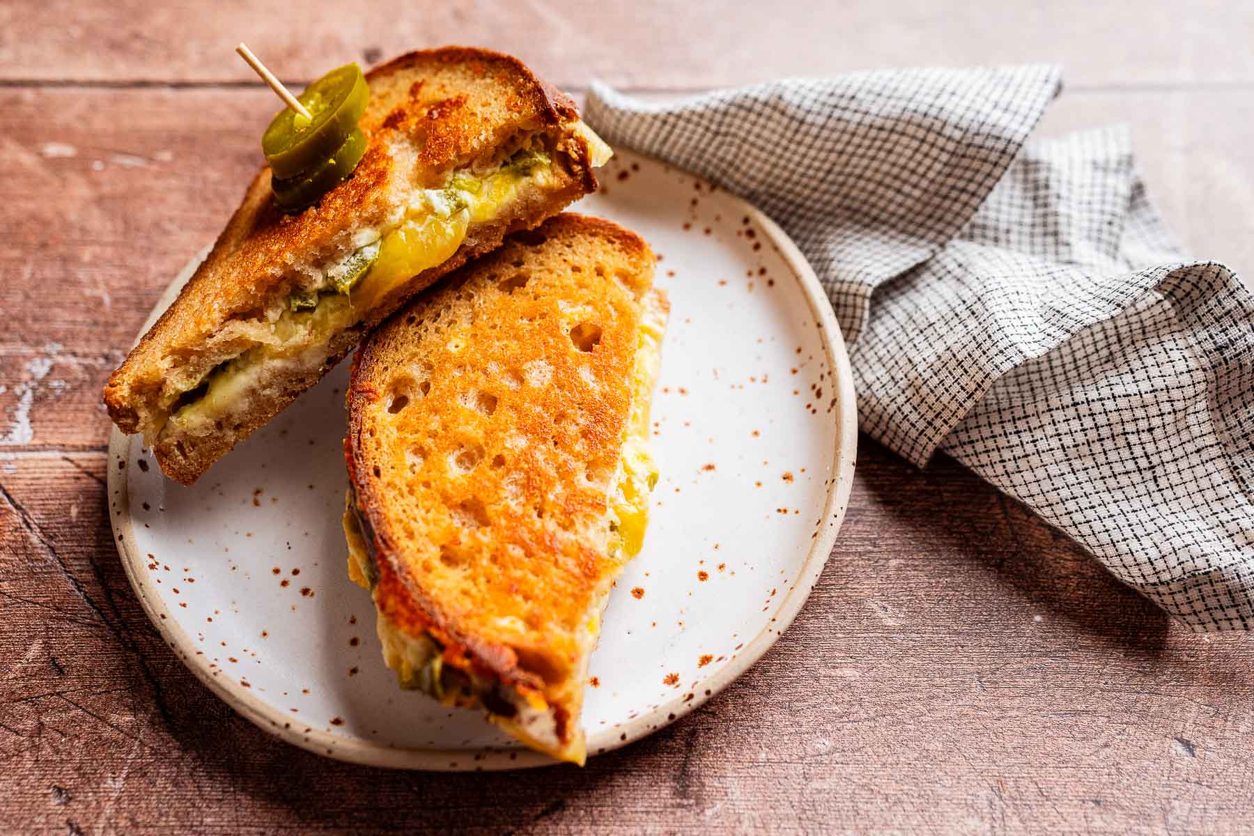 A jalapeno popper grilled cheese sandwich, cut in half and stacked on a white speckled plate, topped with a slice of jalapeño and served beside a checkered cloth.