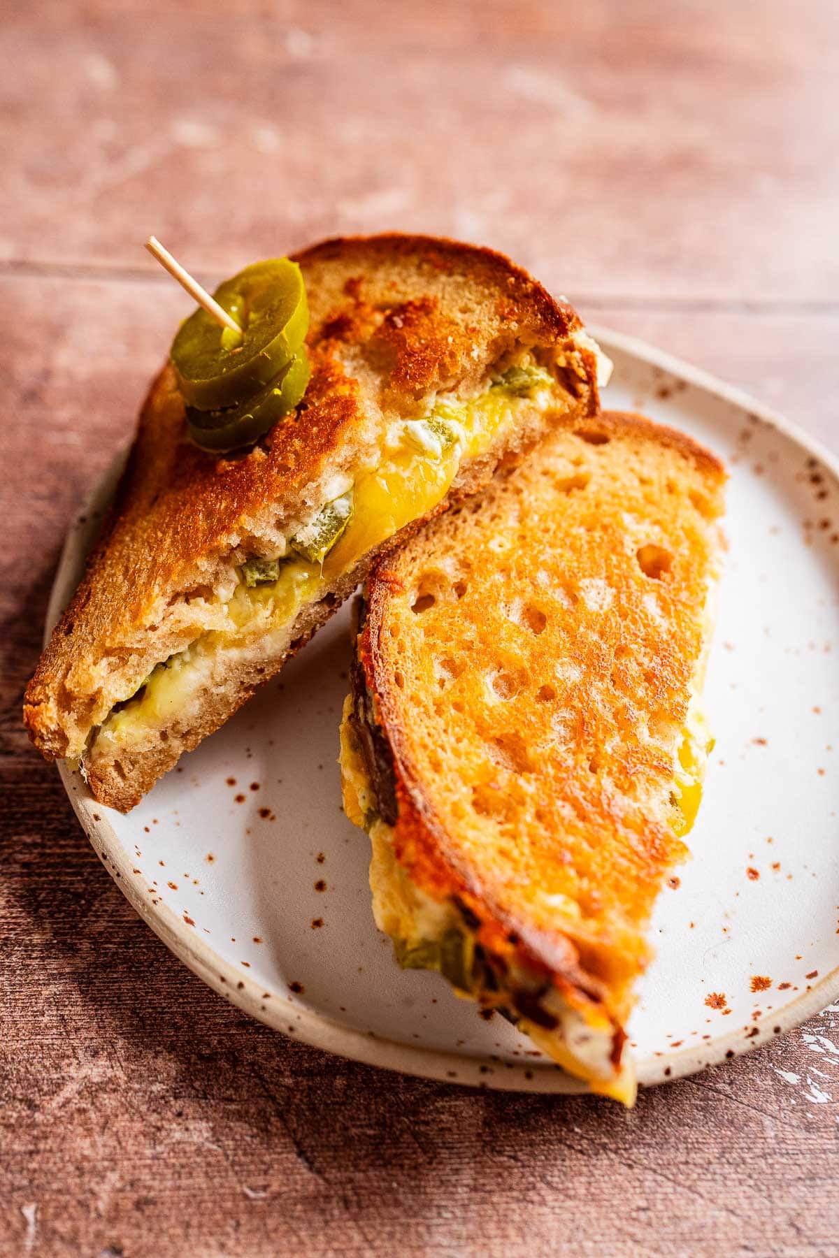 A jalapeno popper grilled cheese sandwich with melted cheese and sliced jalapeños, cut in half and served on a speckled plate.