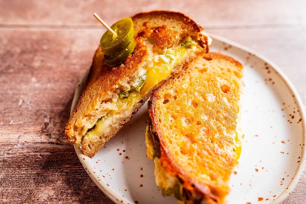 A jalapeno popper grilled cheese sandwich cut in half on a plate, topped with sliced jalapeños and secured with a toothpick.