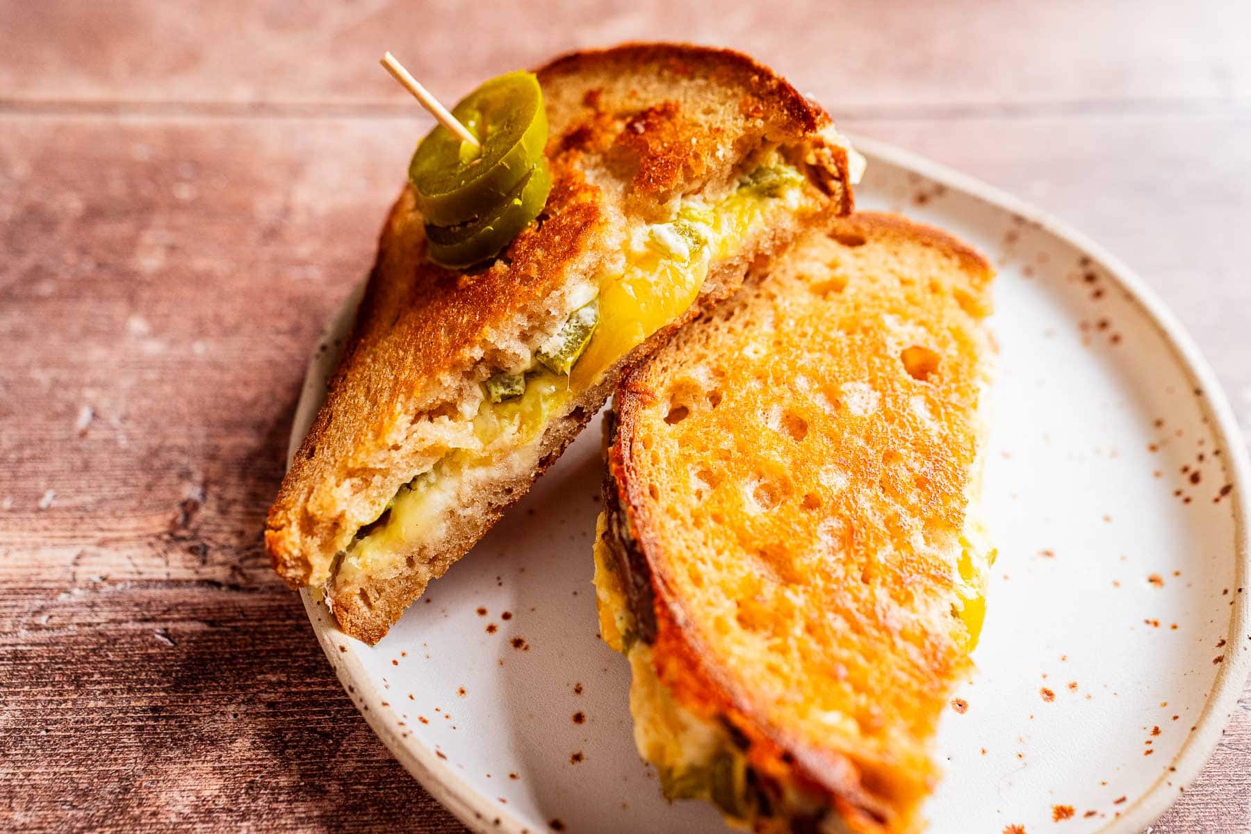 A jalapeno popper grilled cheese sandwich cut in half on a plate, topped with sliced jalapeños and secured with a toothpick.