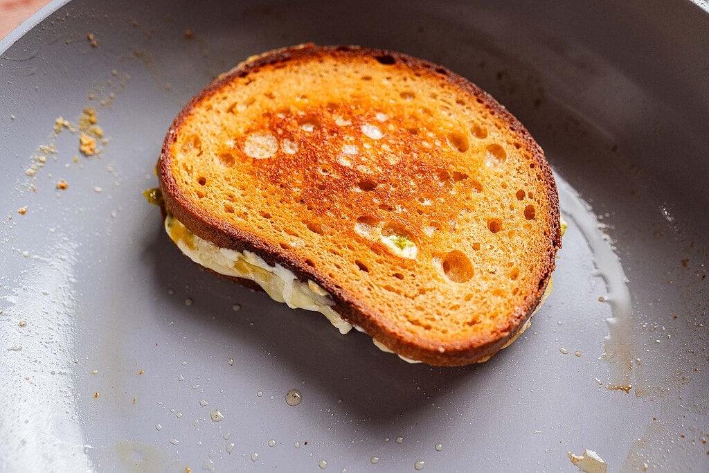 A jalapeno popper grilled cheese sandwich with golden brown, crispy bread sits in a frying pan, hints of spicy peppers and creamy cheese melding with the melted butter.