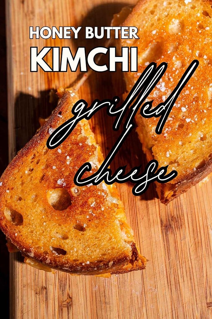 Two halves of a golden-brown kimchi grilled cheese sandwich rest on a wooden surface, with the text "Honey Butter Kimchi Grilled Cheese" overlaid on the image.