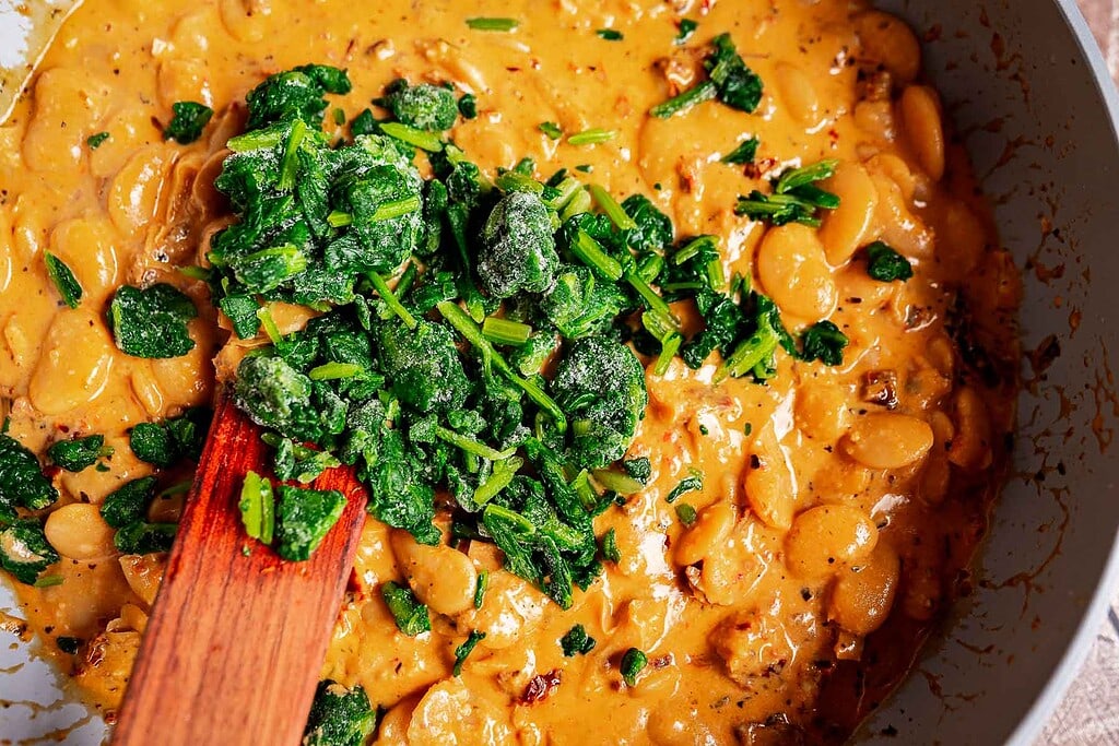 A creamy marry me butter beans mixture is being stirred in a pan, with frozen chopped spinach added on top.