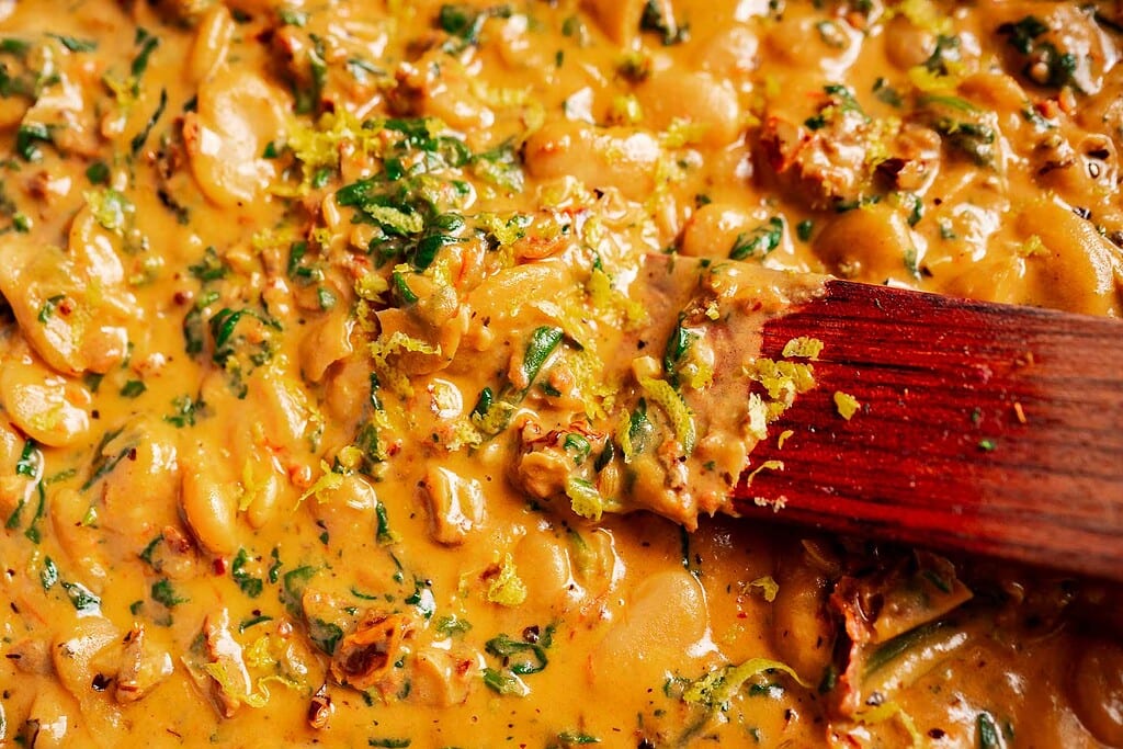 A close-up of creamy marry me butter beans stew with greens and lemon zest, being stirred with a wooden spoon.
