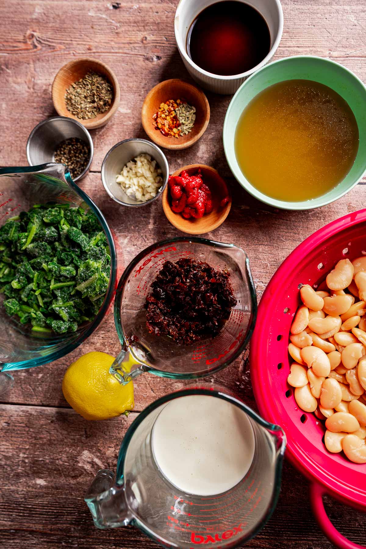 Various ingredients on a wooden table, including beans, chopped greens, lemon, spices, tomato paste, garlic, broth, plant-based milk and a bowl of chopped sun-dried tomatoes for the Mary Me Butter Beans recipe.