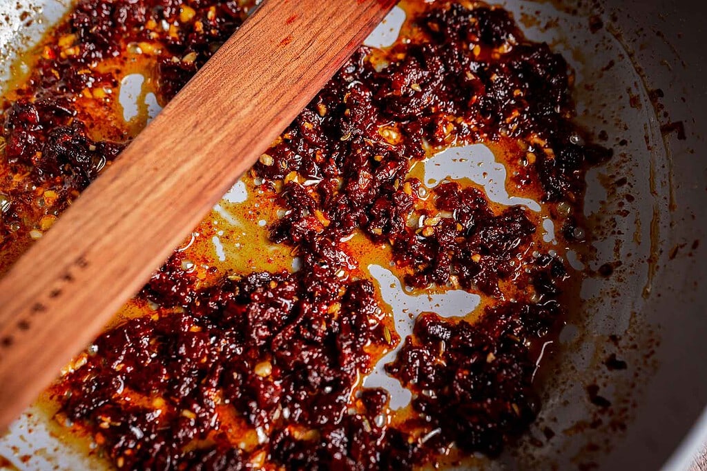 Chopped sun-dried tomatoes and oil are stirred with a wooden spatula in a pan, creating the perfect base for marry me butter beans.