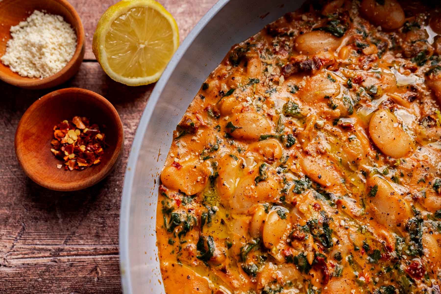 Close-up of a bowl of creamy married butter beans with herbs and spices, small bowl of grated cheese on the side, red pepper flakes and a lemon half on a wooden surface.