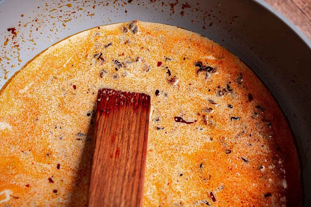 A wooden spoon rests in a pot of creamy, orange-red soup—marry me butter beans add richness, with visible spices and seasoning floating on the surface.