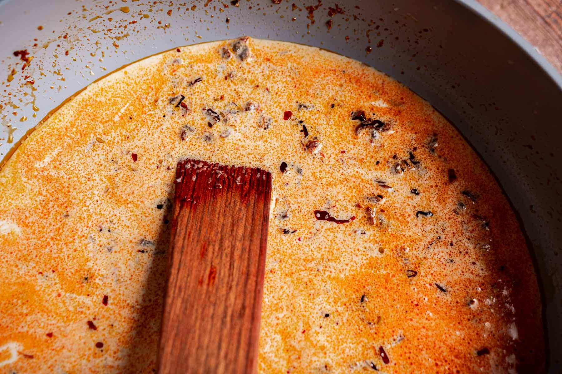 A wooden spoon rests in a bowl of creamy, orange-red soup—Mary Mee butter beans add richness, with visible spices and seasonings floating to the surface.