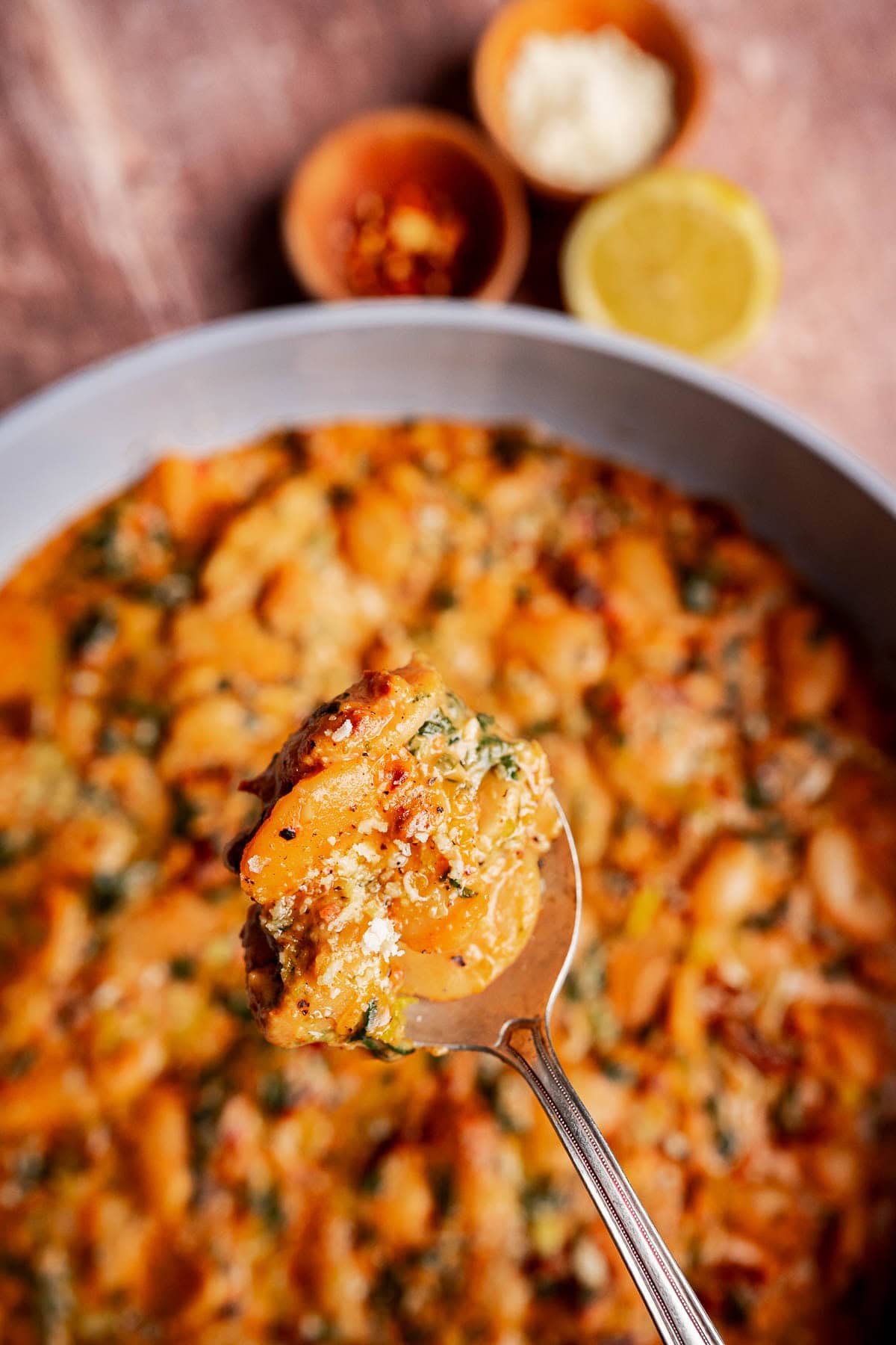 A close-up of a spoon holding cheesy baked gnocchi with herbs on top of a pan in the same dish. Small bowls with Parmesan, red pepper flakes, and lemon halves are in the background—perfect alongside Mary Me Butter Beans.
