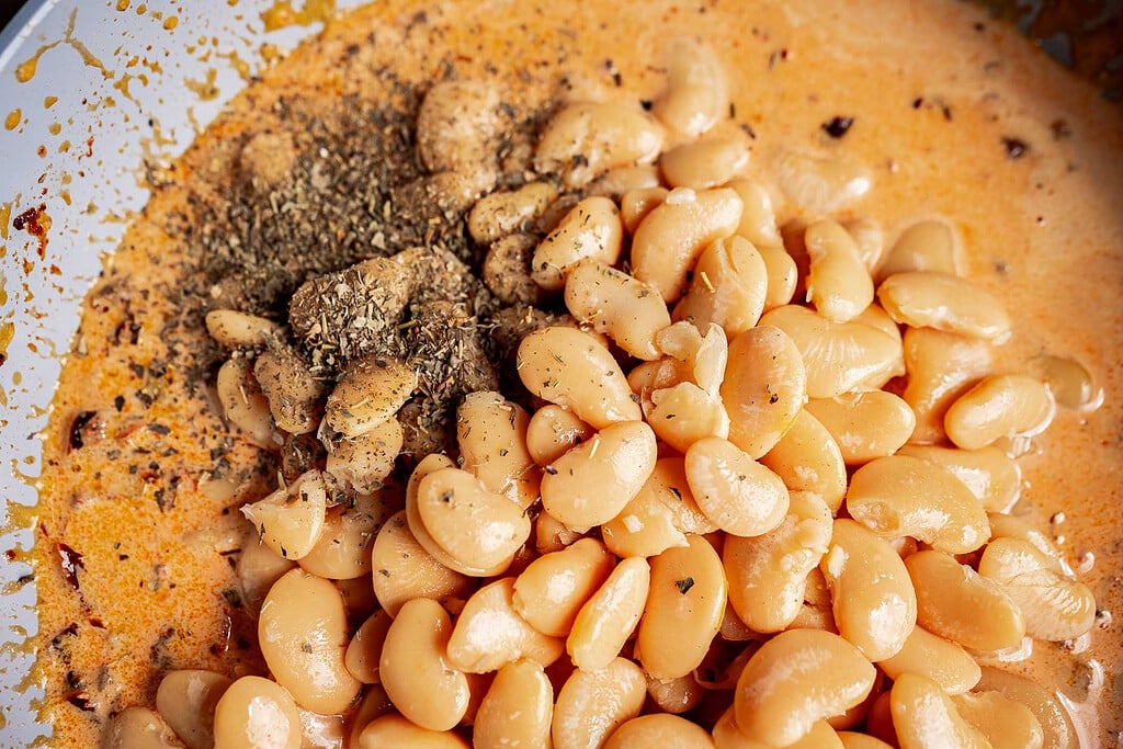 Close-up of large white marry me butter beans in a creamy orange sauce, topped with dried herbs and seasoning.