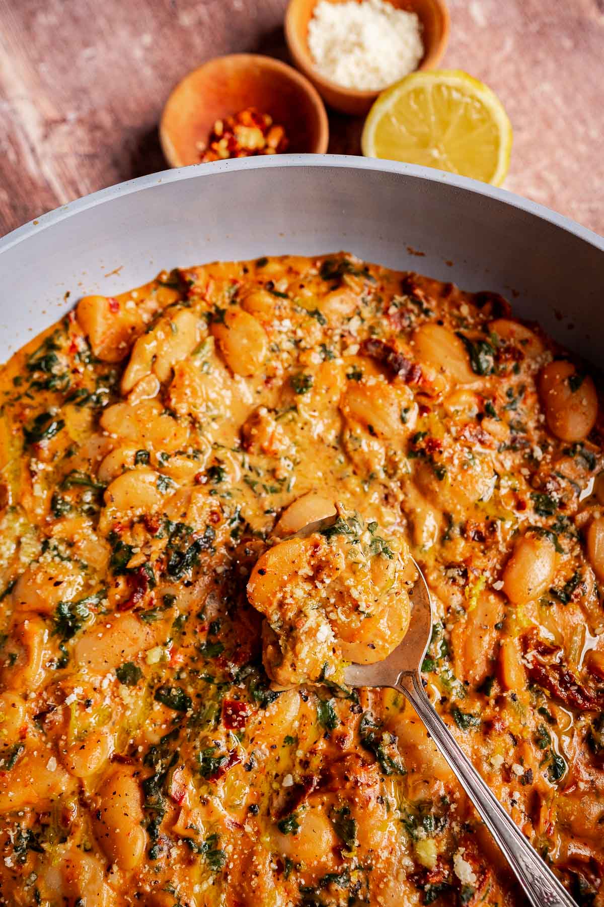 A spoon lifts creamy “marry me butter beans” in a tomato and spinach sauce from a pan, with bowls of grated cheese, red pepper flakes, and a halved lemon in the background.
