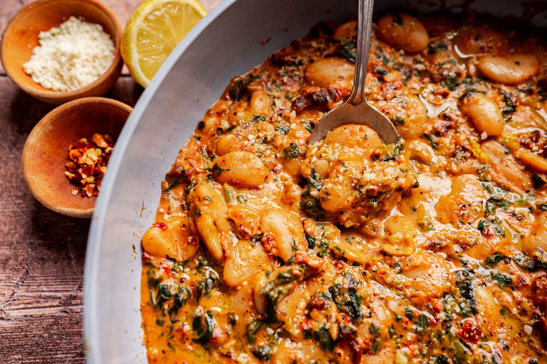 A close-up of a creamy Mary Mee butter beans stew with herbs in a pan, scooping some out with a spoon; Lemon, chili flakes and grated cheese are in small bowls nearby.