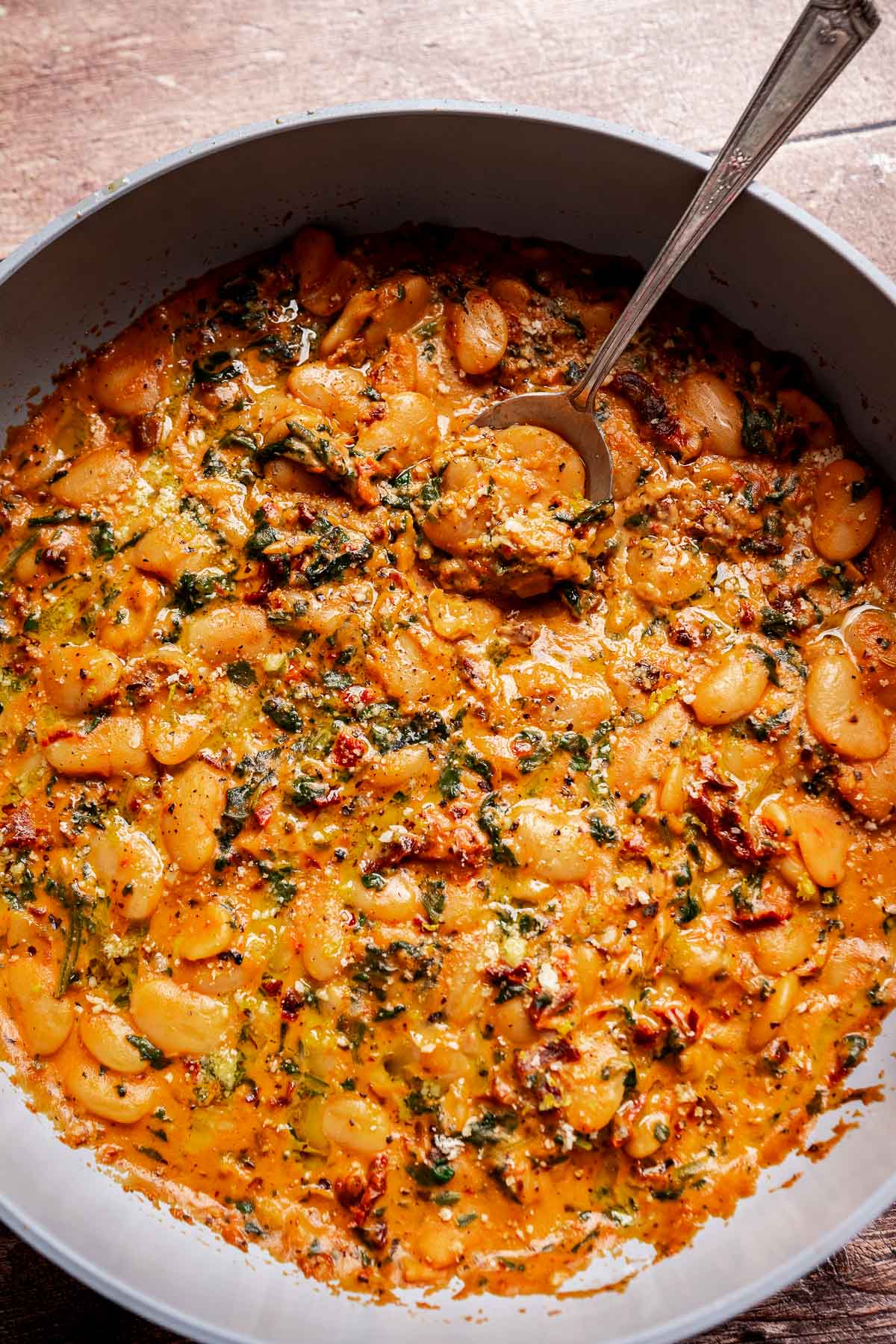 A skillet filled with creamy tomato and herb sauce with large white Mary Mee butter beans, garnished with herbs, and a spoon resting inside.