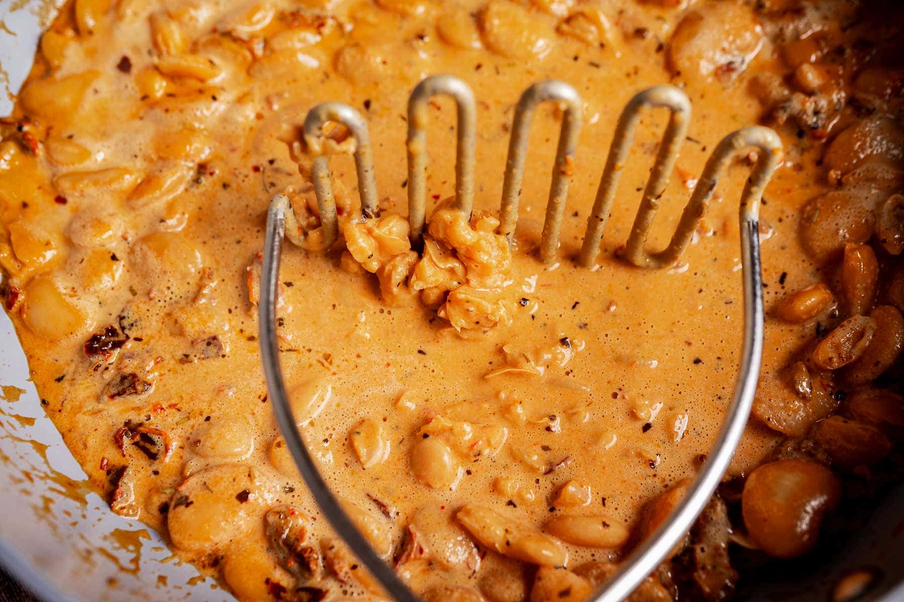A potato masher presses cooked Mary Mee butter beans into a creamy, orange sauce inside a pan.